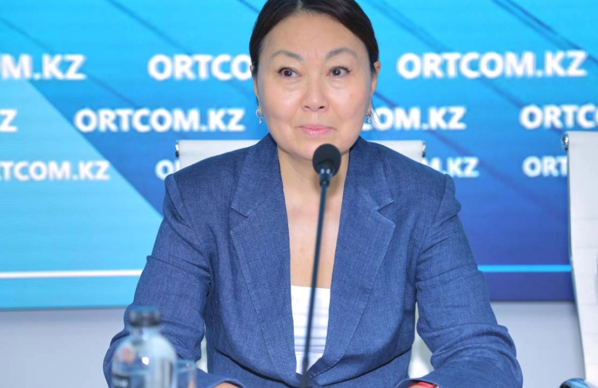 Here's a concise alt text description:

> A woman in a blue blazer sits at a table with a microphone, speaking at a press conference or similar event.  The background features a blue banner with a website address. - МЭКС