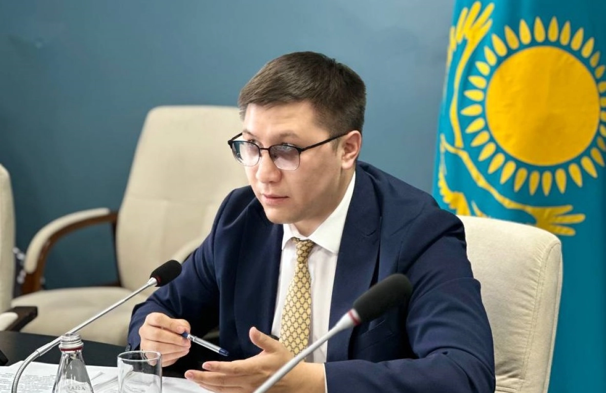Man in suit and glasses speaks at a table with microphones, the Kazakhstani flag visible in the background. - МЭКС