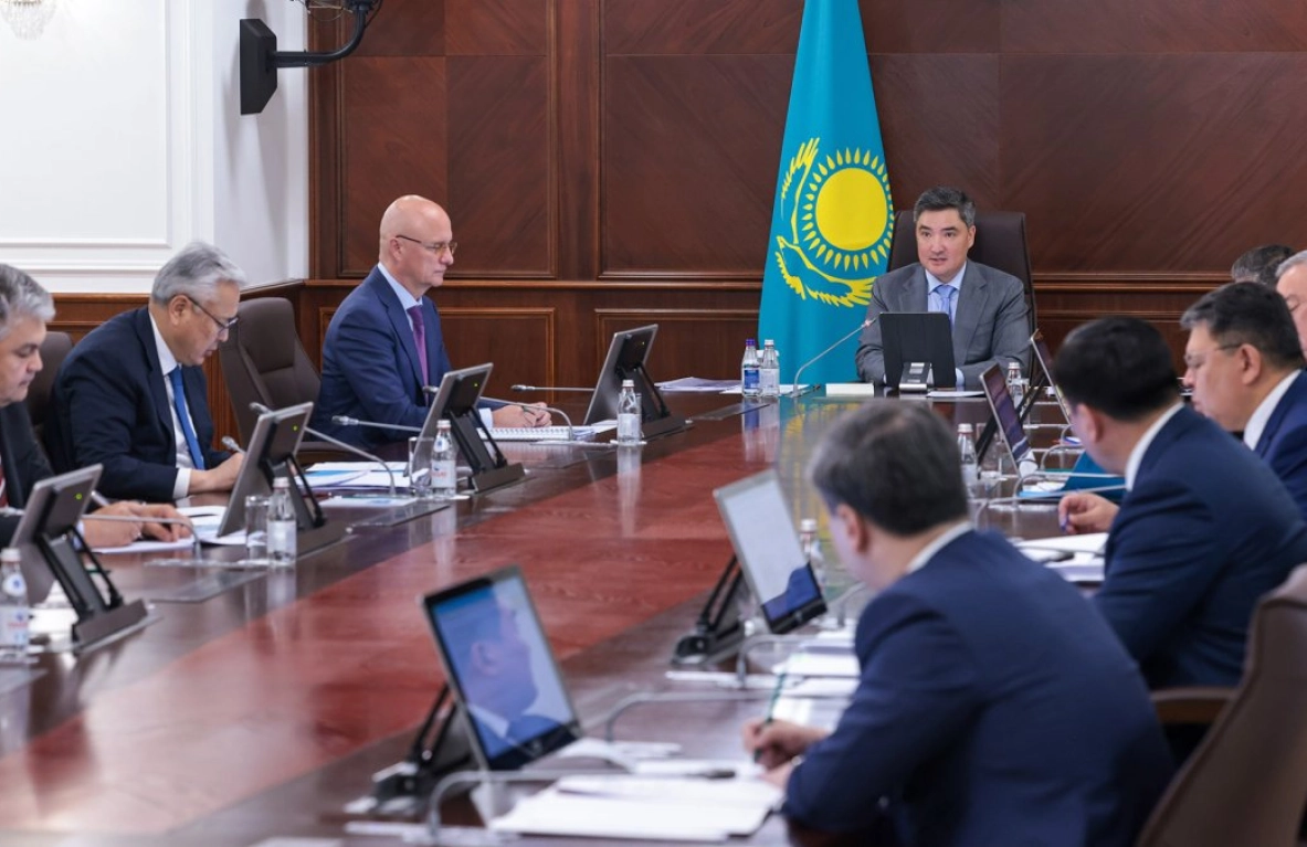 A group of people in suits sit around a large conference table, listening to a speaker at the head of the table.  The Kazakhstani flag is visible in the background. - МЭКС